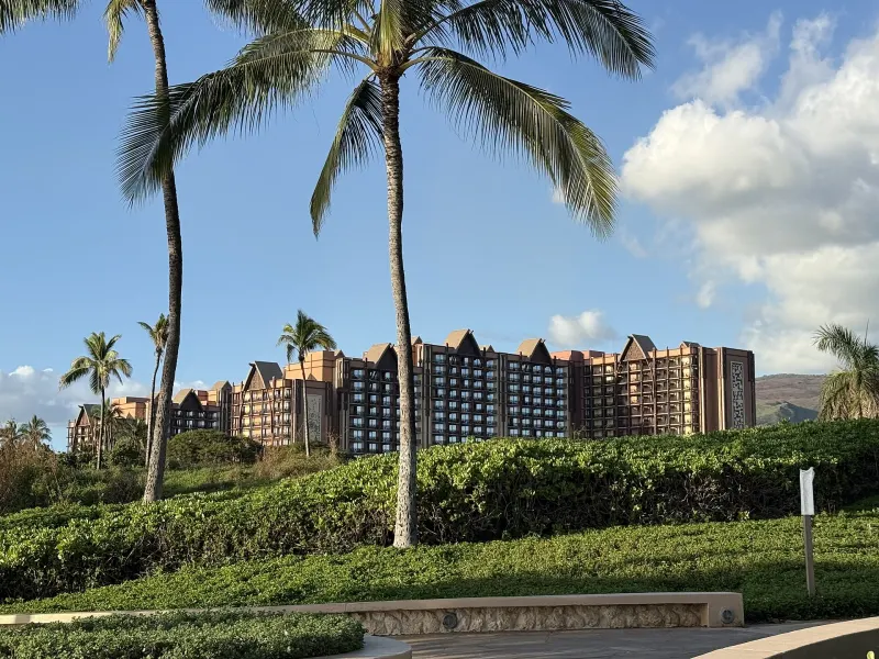 Aulani's resort towers behind tall palm trees and landscaped hedges in afternoon light