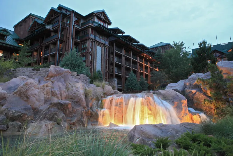Disney's Wilderness Lodge at dusk, with a lit waterfall cascading over rocks below the lodge's timber balconies