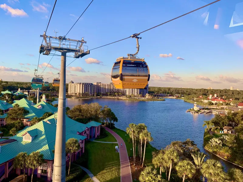 A Disney Skyliner gondola above Barefoot Bay, with Caribbean Beach's green-roofed buildings below and Disney's Riviera Resort across the water